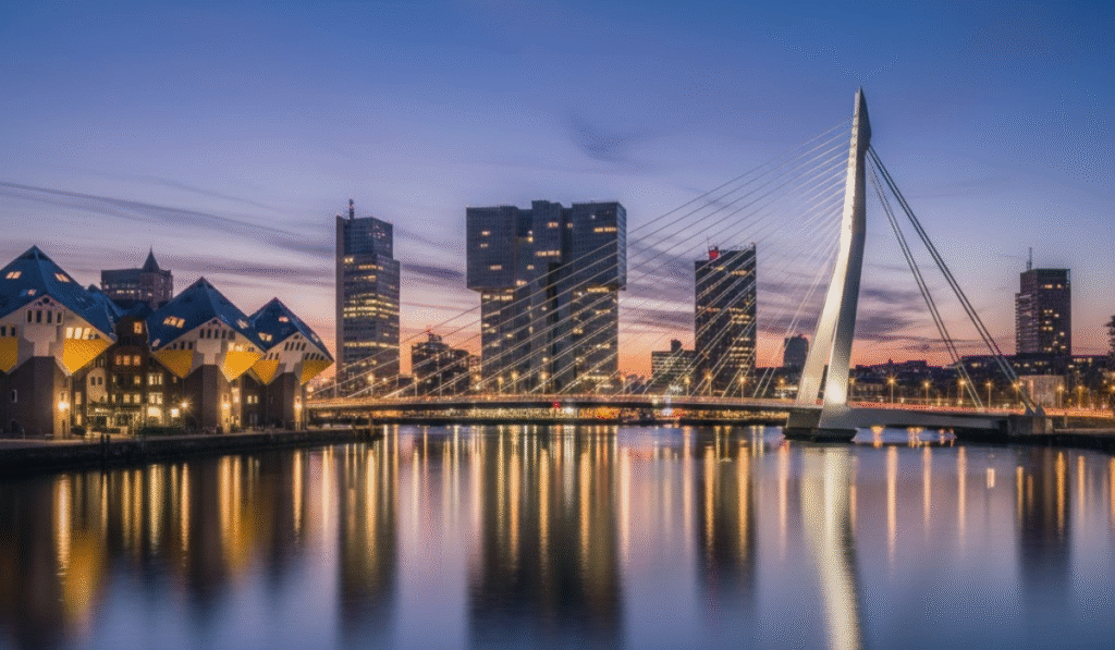 Striking twilight skyline of Rotterdam, Netherlands, featuring the iconic Erasmus Bridge, modern high-rise buildings, and the unique Kubuswoningen (Cube Houses), all reflected in the calm Maas River under a gradient blue and orange sky.

